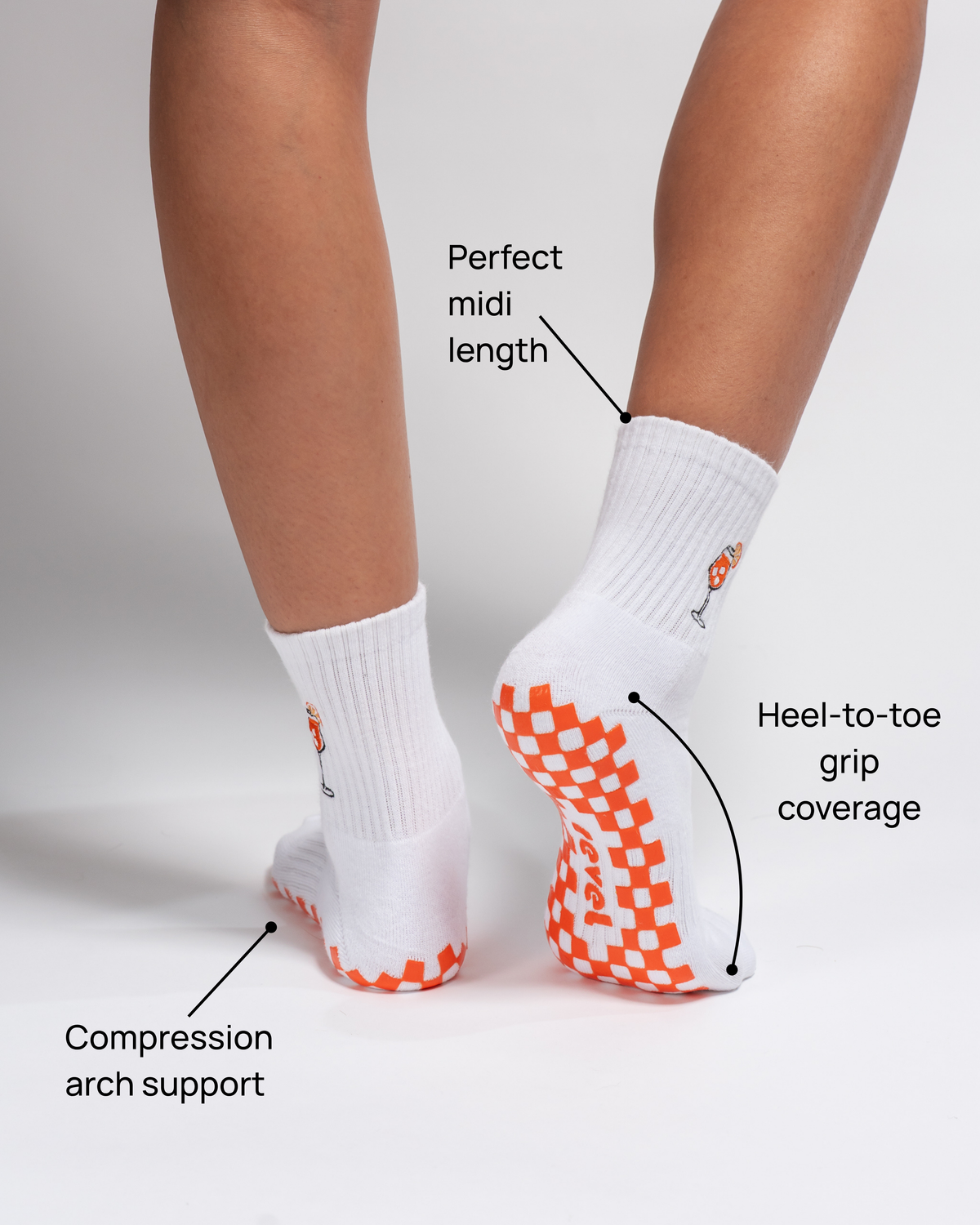 A person is shown from behind wearing The Spritz Grip Sock by Revel Athletic—white crew socks featuring orange checkered grips on the soles. Designed with compression arch support, a perfect midi length, and heel-to-toe grip coverage, these socks are ideal for your next Pilates or barre class. The plain background highlights the sock’s details and fit.