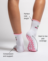 A close-up of feet wearing Revel Athletic’s The Los Angeles Grip Sock—white Pilates grip socks with pink trim and tiny car designs. Labels highlight the midi length, full heel-to-toe grip coverage, and compression arch support.