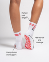 A person wears Revel Athletic’s The New York Grip Sock—white with red accents and non-slip soles—perfect for yoga or Pilates. Text highlights its perfect midi length, full heel-to-toe grip coverage, and compression arch support. The socks read “I ♥ ME.”.
