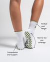 Model is wearing The Extra Dirty Grip Sock by Revel Athletic in white with green trim and checkered grip soles. Text labels highlight features: Perfect midi length, Heel-to-toe grip coverage, and Compression arch support. These Pilates grip socks are designed to deliver stability and comfort throughout your practice. Background is plain white.