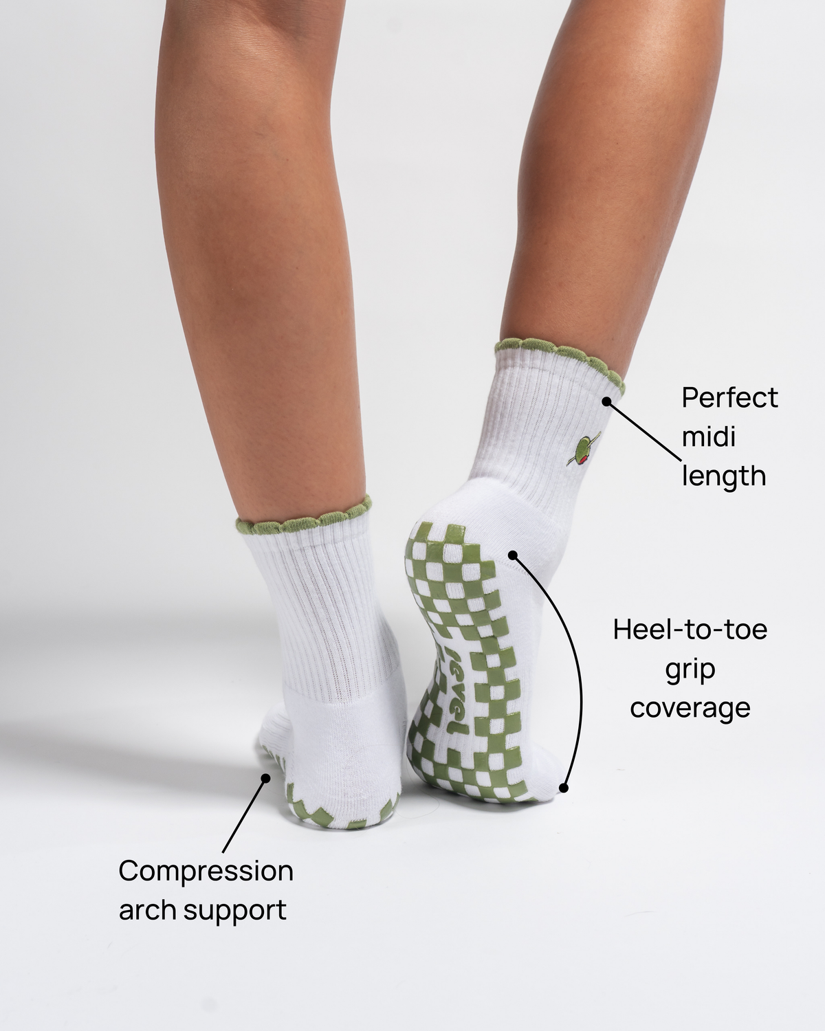 Model is wearing The Extra Dirty Grip Sock by Revel Athletic in white with green trim and checkered grip soles. Text labels highlight features: Perfect midi length, Heel-to-toe grip coverage, and Compression arch support. These Pilates grip socks are designed to deliver stability and comfort throughout your practice. Background is plain white.