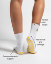 A person wearing The I’m Fine Grip Sock by Revel Athletic in white, featuring a midi length and yellow checkered grip soles. Text highlights heel-to-toe grip coverage, compression arch support, and the ideal midi length.