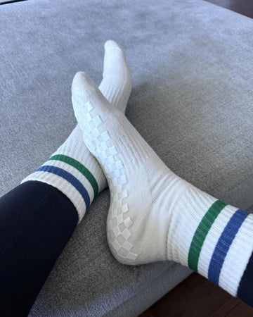 Person wearing white pilates socks with green and blue stripes sits with legs crossed on a light gray ottoman. The grippy socks have textured grips on the soles. Part of dark pants and wooden floor are visible.