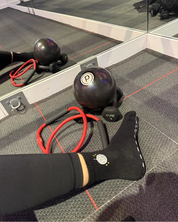 A person’s leg in black leggings and grippy socks stretches out on a gym floor near a weighted ball, resistance band, and small exercise ball in front of mirrors. The grippy socks provide extra stability during the workout.