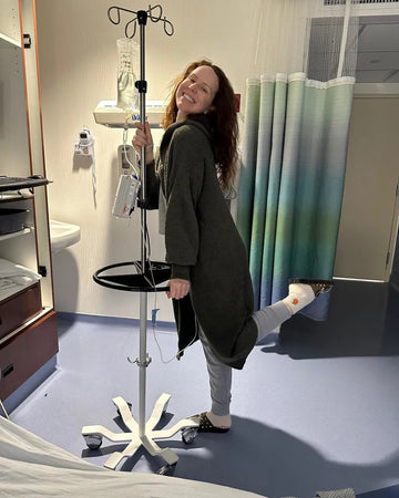 A woman in a hospital room smiles while holding an IV stand. She wears a long robe, has one leg raised, and has a bandage on her foot with a pair of sticky socks for added comfort and safety. Hospital equipment and a curtained area are visible in the background.