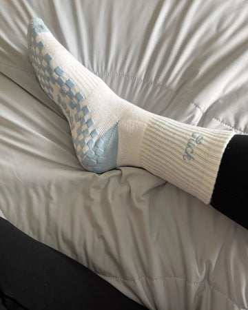 A person wearing white pilates socks with light blue grip patterns and black pants rests their foot on a light gray comforter.