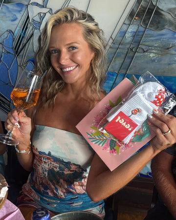 Smiling woman with wavy blonde hair holds a glass of orange drink in one hand and a pink gift bag with a card and baby grip socks in the other, sitting at a table in a restaurant.