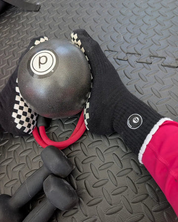A person wearing black gloves with checkered cuffs and barre socks holds a weighted exercise ball with a P logo; dumbbells and a red resistance band are on a textured gym mat.