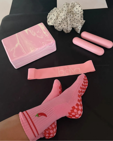 Person wearing pink grip socks with red chili and checkerboard pattern, resting feet on a black surface. Nearby are pink exercise cards, a scrunchie, two pink Bala bars, and a pink Bala resistance band.