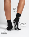 A person wears Revel Athletic’s The All Nighter Grip Sock in black, featuring white checkered soles—perfect for Yoga, Barre, or Pilates. Text highlights include: ideal midi length, full heel-to-toe grip coverage, and supportive compression arch. The background is plain white.