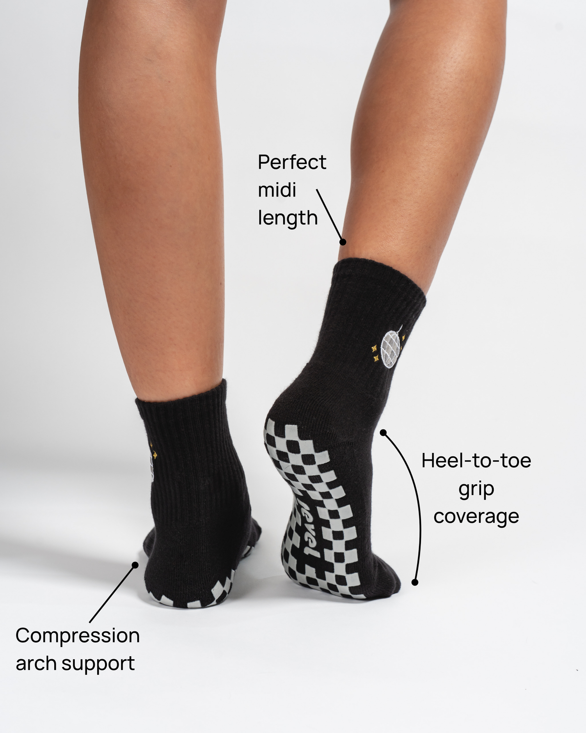 A person wears Revel Athletic’s The All Nighter Grip Sock in black, featuring white checkered soles—perfect for Yoga, Barre, or Pilates. Text highlights include: ideal midi length, full heel-to-toe grip coverage, and supportive compression arch. The background is plain white.