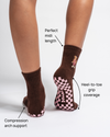 A person wears Revel Athletic’s The Teddy Grip Sock in brown, featuring a pink checkerboard grip on the soles. Arrows highlight features: compression arch support, perfect midi length, and heel-to-toe grip coverage.