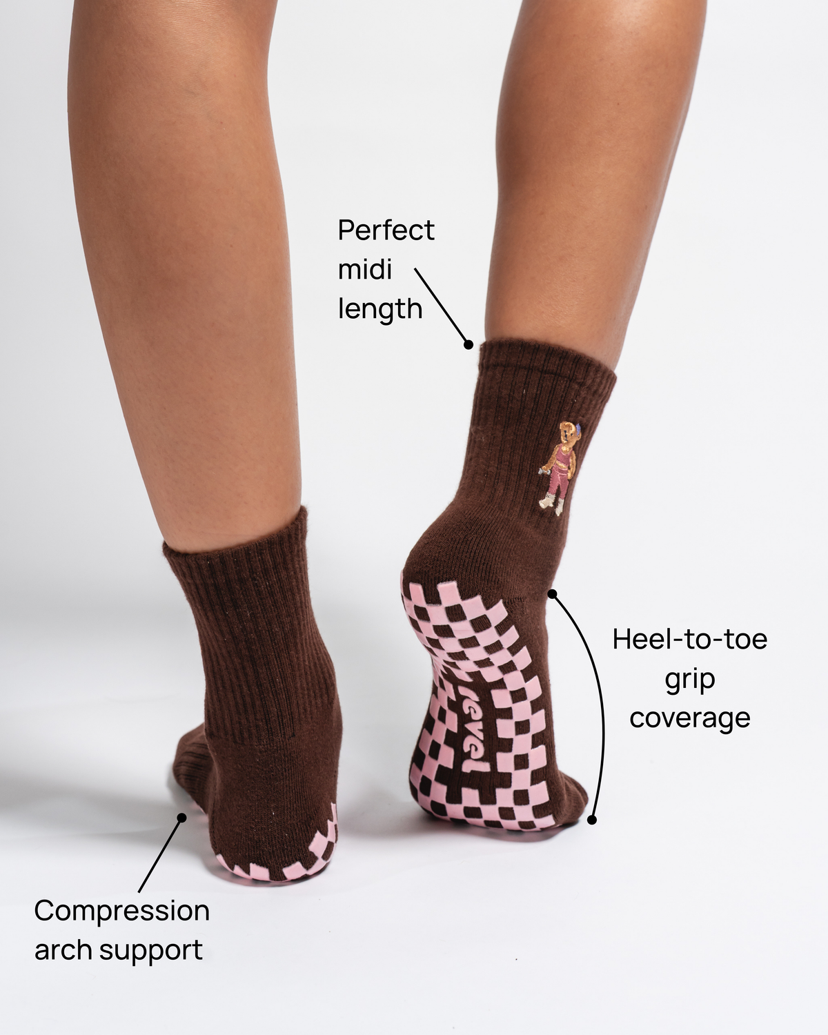 A person wears Revel Athletic’s The Teddy Grip Sock in brown, featuring a pink checkerboard grip on the soles. Arrows highlight features: compression arch support, perfect midi length, and heel-to-toe grip coverage.