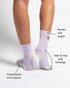 A person stands on a white background wearing The Austin Grip Sock by Revel Athletic in light purple, featuring perfect midi length, heel-to-toe non-slip grip soles—ideal for barre or Pilates—and compression arch support.