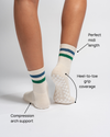 A person wears The Varsity Grip Sock by Revel in a cream color with green and blue stripes. Text highlights key features: compression arch support, heel-to-toe grip coverage, and a perfect midi length. Set against a plain white background, these ideal Pilates socks are designed by Revel for exceptional comfort and stability through every movement.