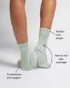 A person wears The Tied & True Grip Sock by Revel Athletic in light green with white bows, highlighting features such as the perfect midi length, heel-to-toe grip coverage for enhanced stability during Yoga and Pilates, and compression arch support. The back view reveals a stylish white checker pattern on the soles.