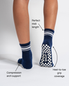 A person wears Revel Athletic’s The Country Club Grip Sock in navy, featuring white stripes and checkered grip soles—perfect for Pilates or barre classes. The midi-length socks offer compression arch support and heel-to-toe grip coverage. Only the lower legs and feet are visible.