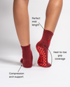Two feet wearing The Very Cherry Grip Sock by Revel Athletic are shown, with labels highlighting compression arch support, a perfect midi length, and heel-to-toe grip coverage. Ideal as barre yoga socks or Pilates grip socks, these stylish maroon socks with cherry embroidery stand out against a crisp white background.