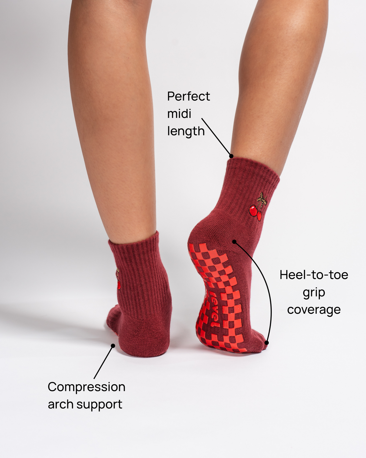 Two feet wearing The Very Cherry Grip Sock by Revel Athletic are shown, with labels highlighting compression arch support, a perfect midi length, and heel-to-toe grip coverage. Ideal as barre yoga socks or Pilates grip socks, these stylish maroon socks with cherry embroidery stand out against a crisp white background.