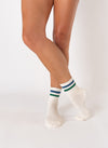 A person stands on one leg with bare legs crossed, showcasing Revel's The Varsity Grip Sock in white, featuring blue and green stripes at the top, against a plain white background.