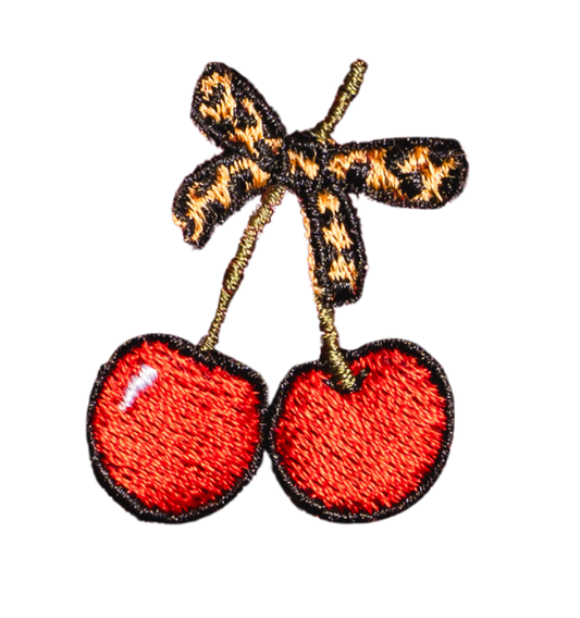 Embroidered patch of two red cherries with green stems tied together by a small bow featuring a yellow and black leopard print pattern. Perfect for personalizing your favorite accessories, like grippy socks or backpacks, the design is outlined in black on a white background.
