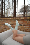 A person relaxes on a light gray couch with legs raised, wearing Revel Athletic's The Tied & True Grip Sock in mint green and beige shorts. Behind them, brick walls and large rustic windows frame the view of bare trees outside.