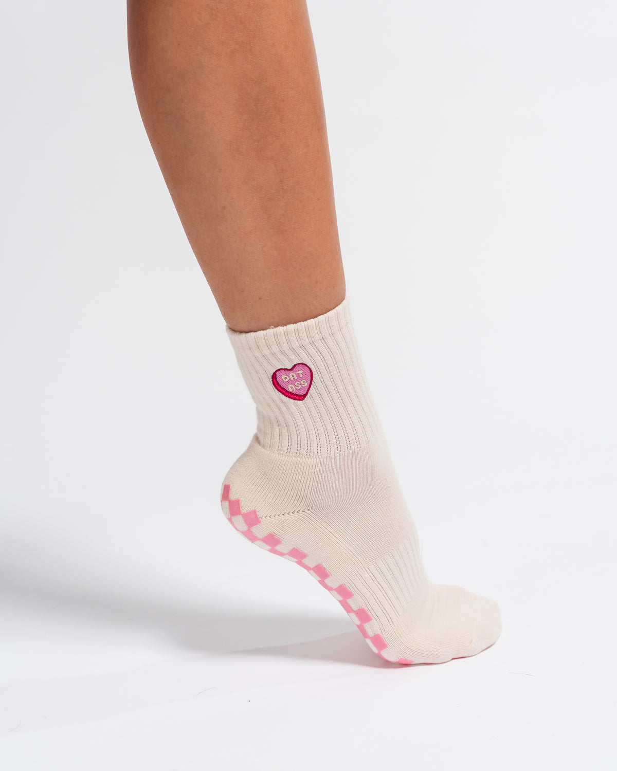 A person wearing Revel Athletic’s The Sweet Nothings Grip Sock—a cream sock featuring pink zigzag grip patterns on the sole and a pink heart patch on the side—stands on a white background.
