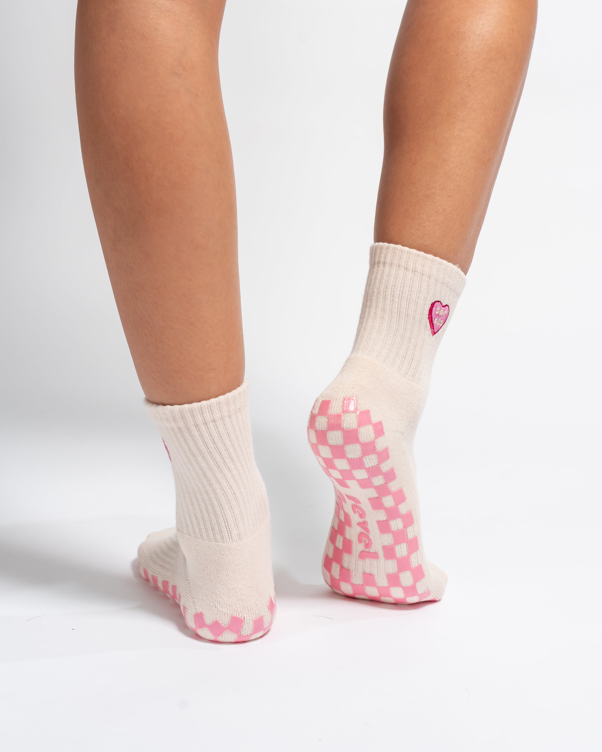 Person wearing cream-colored grippy socks with pink checkered soles and a small pink heart on the back of each ankle, standing on a white background. These pilates socks are perfect for maintaining stability and style during your workout.