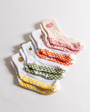 Five pairs of socks lay in a row on a white surface. Four pairs are white with different colored checkerboard patterns and fruit or smiley designs, each featuring grippy soles for added comfort. One pair is pink with a heart patch, a pink checkerboard pattern, and sticky grip on the bottom—perfect for those who love both style and the secure fit of grippy socks.