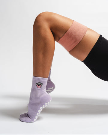 A person wearing a pink resistance band above their knees, black shorts, and light purple pilates socks with a smiling face design, performing a glute bridge exercise on a white background.