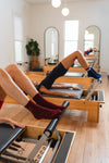 Three people wearing Revel Athletic's The Very Cherry Grip Sock exercise on reformer machines in a bright studio with wooden floors, large mirrors, and a potted plant. They are performing a leg press move with their hips raised.
