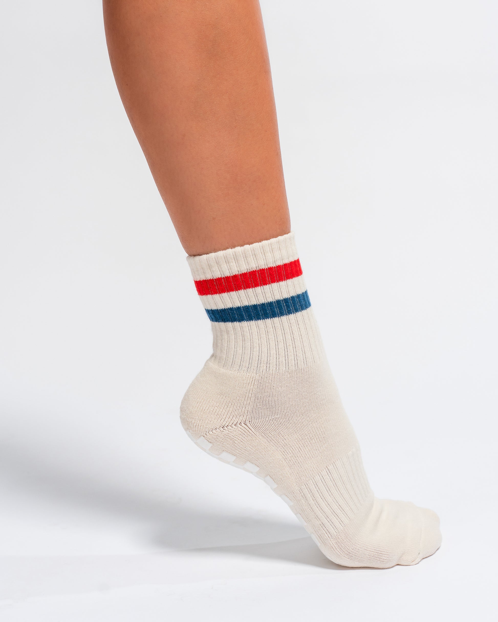 A person wearing The Miss Americana Grip Sock by Revel Athletic, featuring red and blue stripes near the top on an off-white base, stands on a white background.