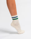 A person wears The Varsity Grip Sock by Revel in a cream color with blue and green stripes and grip pads on the sole, standing on tiptoe against a white background.