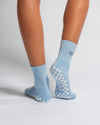 A person is wearing The Cross My Heart Grip Sock by Revel Athletic in light blue, featuring a checkered non-slip pattern on the soles and a small heart design on the back, while standing on a white background.