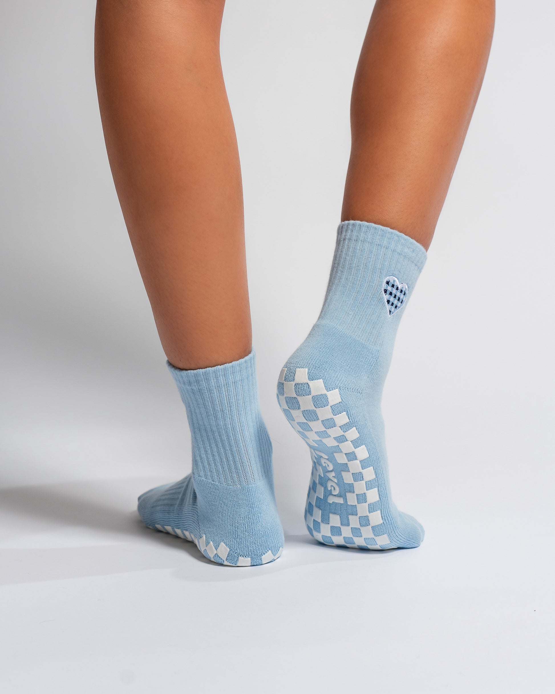 A person is wearing The Cross My Heart Grip Sock by Revel Athletic in light blue, featuring a checkered non-slip pattern on the soles and a small heart design on the back, while standing on a white background.