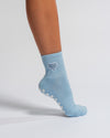 A person’s lower leg wearing The Cross My Heart Grip Sock by Revel Athletic in light blue, featuring a small white and blue checkered heart patch and white non-slip grips on the sole, against a plain white background.