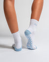 A person is wearing Revel Athletic's The Pardon My French Grip Sock in Baby Blue. These white crew socks have blue heels and soles, a checkered blue grip pattern on the bottom for enhanced stability during yoga or Pilates, and light blue text on the back. The individual is standing on a white surface.
