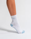 A person wearing The Pardon My French Grip Sock (Baby Blue) by Revel Athletic, featuring white fabric with light blue accents and FLOE text, stands on tiptoe against a plain white background—showcasing the ideal style for Pilates or yoga grip socks.