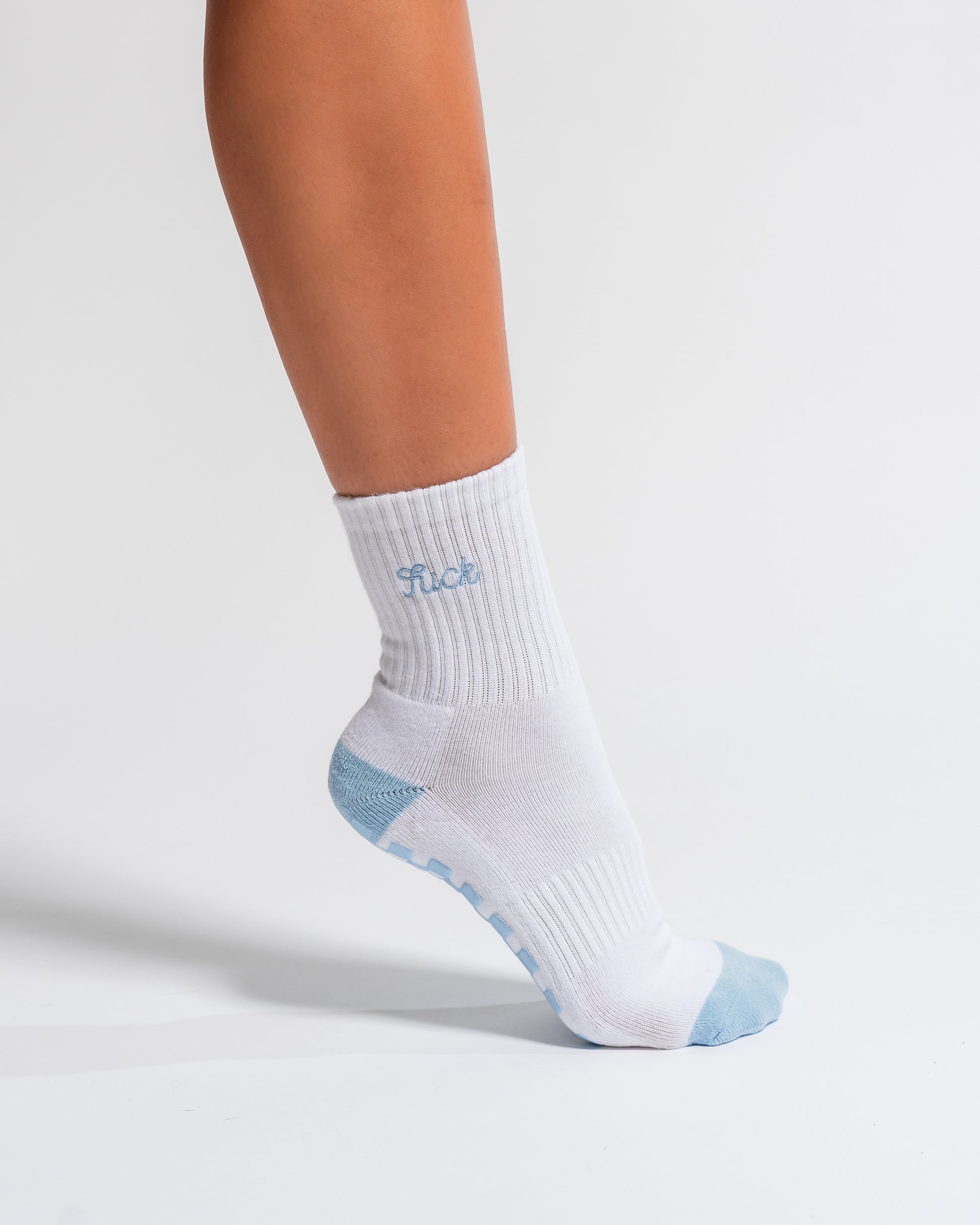 A person wearing The Pardon My French Grip Sock (Baby Blue) by Revel Athletic, featuring white fabric with light blue accents and FLOE text, stands on tiptoe against a plain white background—showcasing the ideal style for Pilates or yoga grip socks.