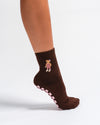 A person wearing The Teddy Grip Sock by Revel Athletic—with a brown color, adorable teddy embroidery, and a pink grippy checker bottom—stands against a plain white background.