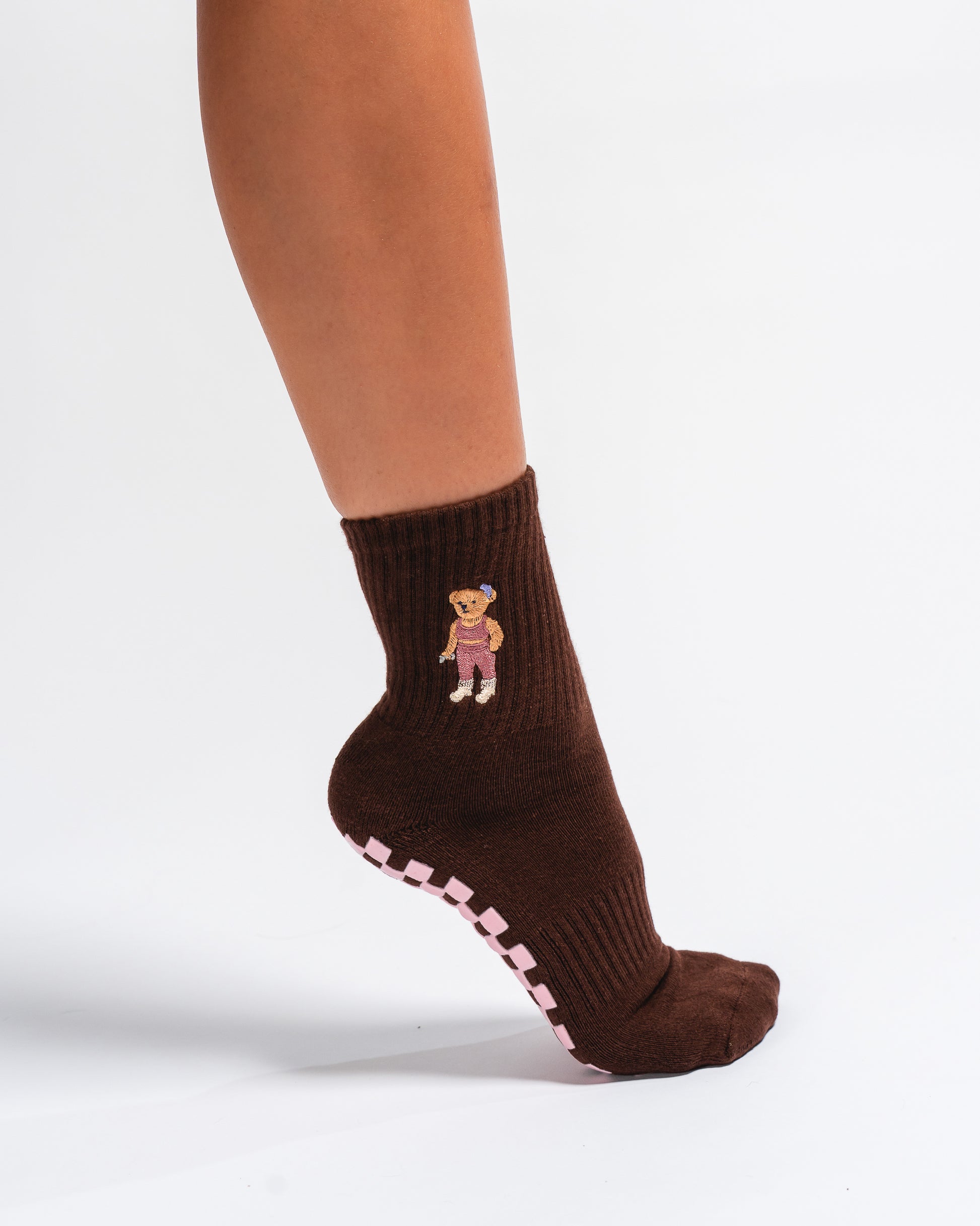 A person wearing The Teddy Grip Sock by Revel Athletic—with a brown color, adorable teddy embroidery, and a pink grippy checker bottom—stands against a plain white background.