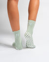 Two legs wearing Revel Athletic’s The Tied & True Grip Sock in light green, featuring small pastel blue bows and a white checkerboard grip pattern on the soles. The background is plain white.