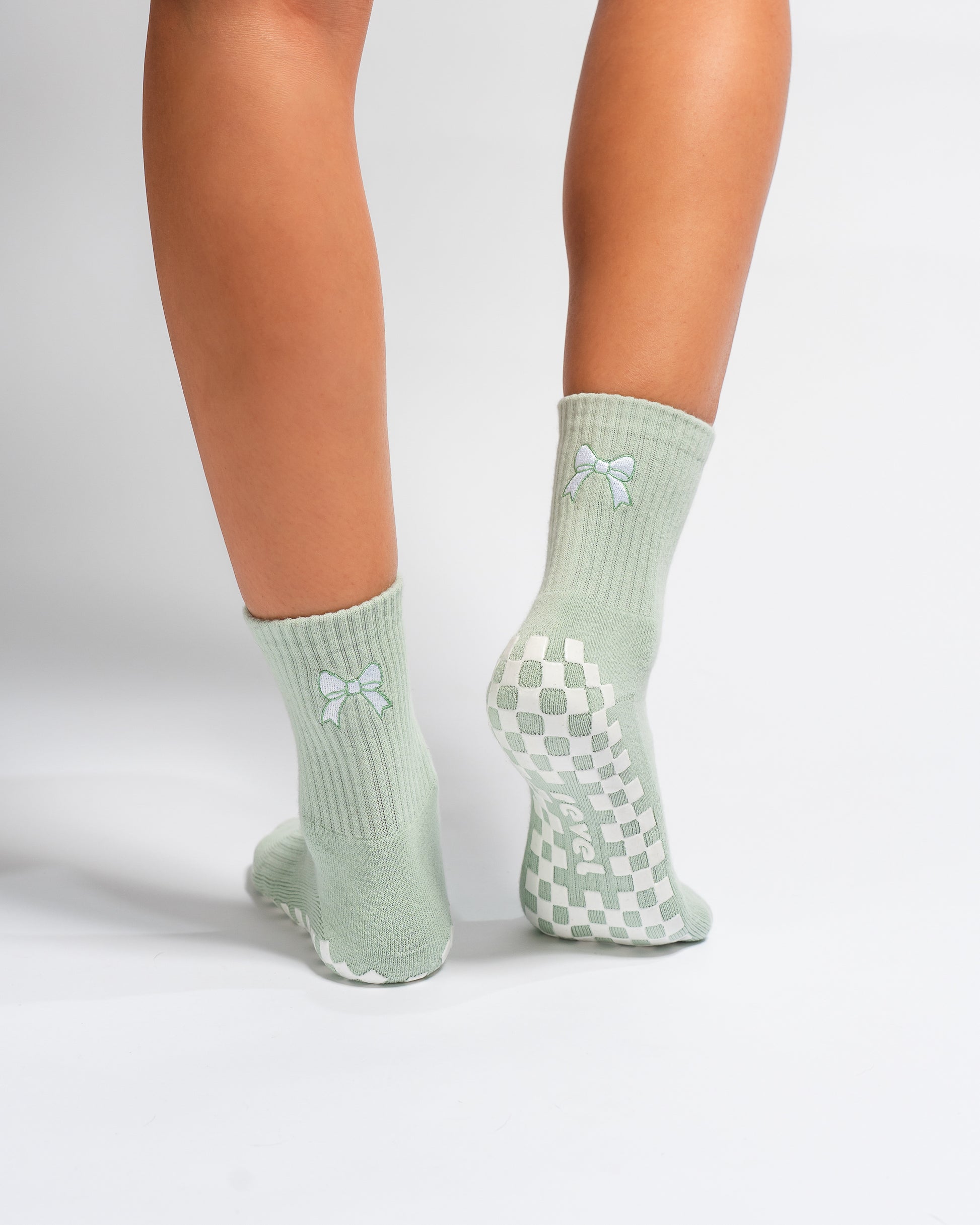 Two legs wearing Revel Athletic’s The Tied & True Grip Sock in light green, featuring small pastel blue bows and a white checkerboard grip pattern on the soles. The background is plain white.