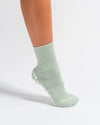 A person wearing the Revel Athletic Tied & True Grip Sock in light green, featuring white non-slip grips on the sole, stands on a white background. Only the lower leg and foot are visible.