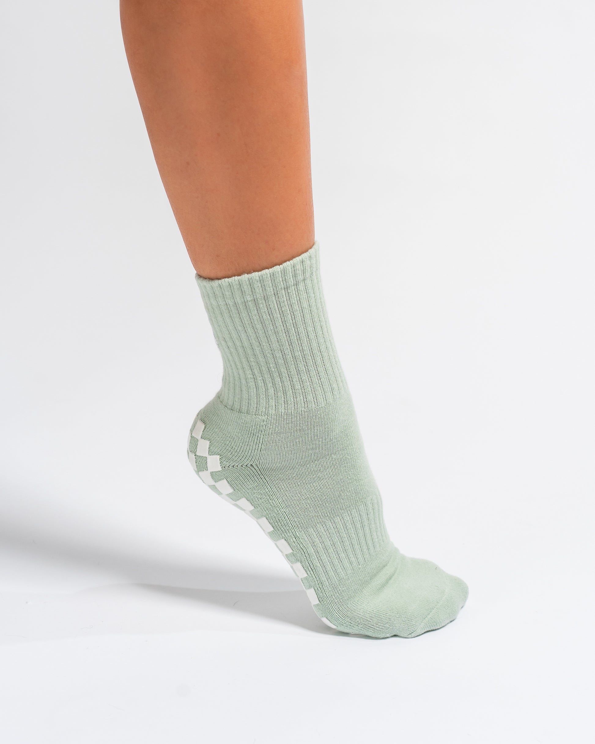 A person wearing the Revel Athletic Tied & True Grip Sock in light green, featuring white non-slip grips on the sole, stands on a white background. Only the lower leg and foot are visible.