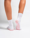 A person wearing The Pardon My French Grip Sock (Petal Pink) by Revel Athletic, featuring ribbed cuffs and pink non-slip grips with text near the top, stands on a white surface.