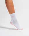 A person wearing Revel Athletic’s The Pardon My French Grip Sock in Petal Pink stands on a white background. This white yoga grip sock features pink accents, a pink logo, ribbed cuffs, and textured grips on the sole—perfect for added stability during your yoga or barre sessions.