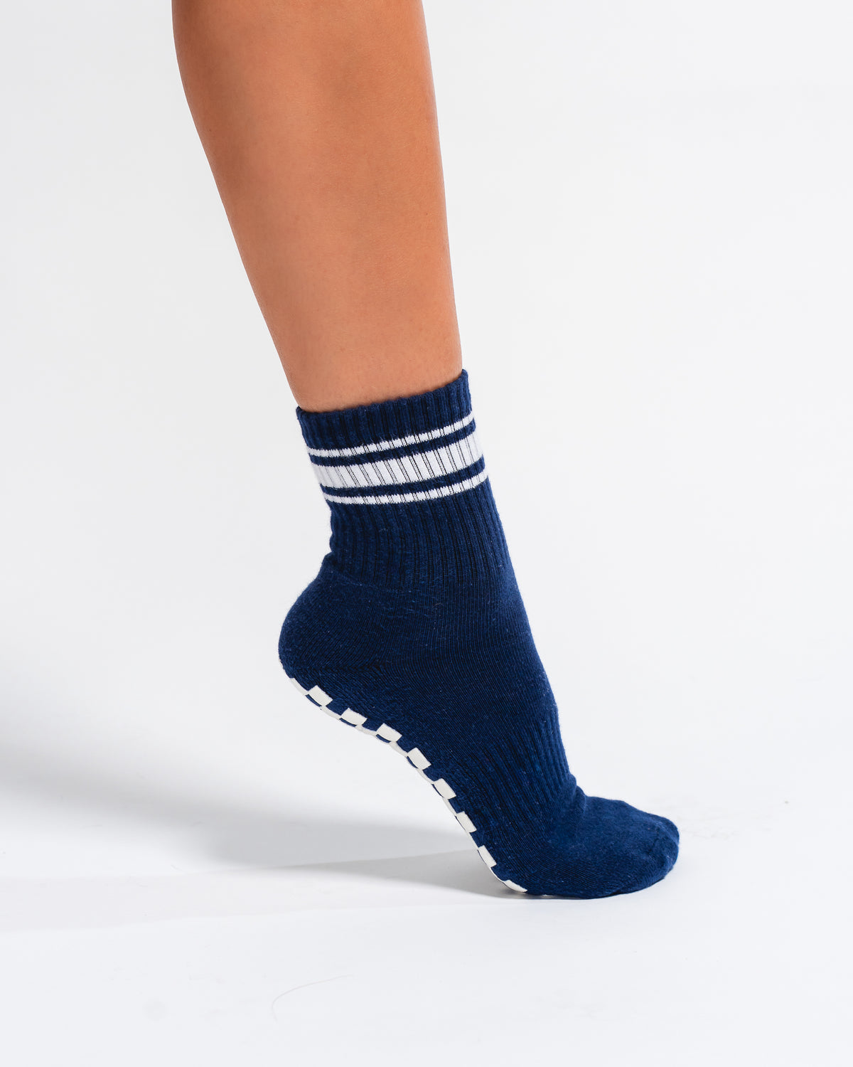 A person wearing The Country Club Grip Sock by Revel Athletic—featuring a navy blue color with white stripes near the ankle and white grip dots on the sole—standing on a white background.