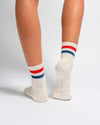 A person stands on tiptoes wearing The Miss Americana Grip Sock by Revel Athletic—cream pilates socks with red and blue stripes. These non-slip socks feature textured, checkerboard grip soles. The background is plain white.