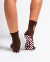 A person wearing Revel Athletic's The Teddy Grip Sock—a brown crew sock with a small teddy embroidery on the side and a pink grippy checker bottom—stands on a white background.